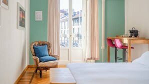 Een kamer met een bed, een stoel, een bureau en uitzicht op gebouwen door een raam.