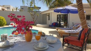 A poolside dining area with a table set for two, a vase of pink flowers, and lounge chairs. - Amour Sénégal (Ngaparou)