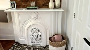 Une cheminée blanche ornée de moulures décoratives, un mur en briques au-dessus, et une étagère blanche sur laquelle sont posés des livres et des vases.