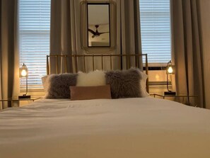 A neatly made bed with fluffy pillows, a ceiling fan, and two bedside lamps in a room with large windows and curtains. - Mulberry Mansion - OTR - FREE Parking (Cincinnati)