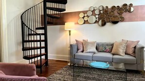Un salon avec un canapé gris, un fauteuil rose, un escalier en colimaçon noir et un mur orné de miroirs ronds.