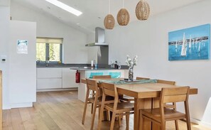 A modern kitchen with a central island, wooden dining table with chairs, and a painting of sailboats on the wall. - Stylish Estuary View | Sleeps 6 | Parking & Balconies (Saint Ives)