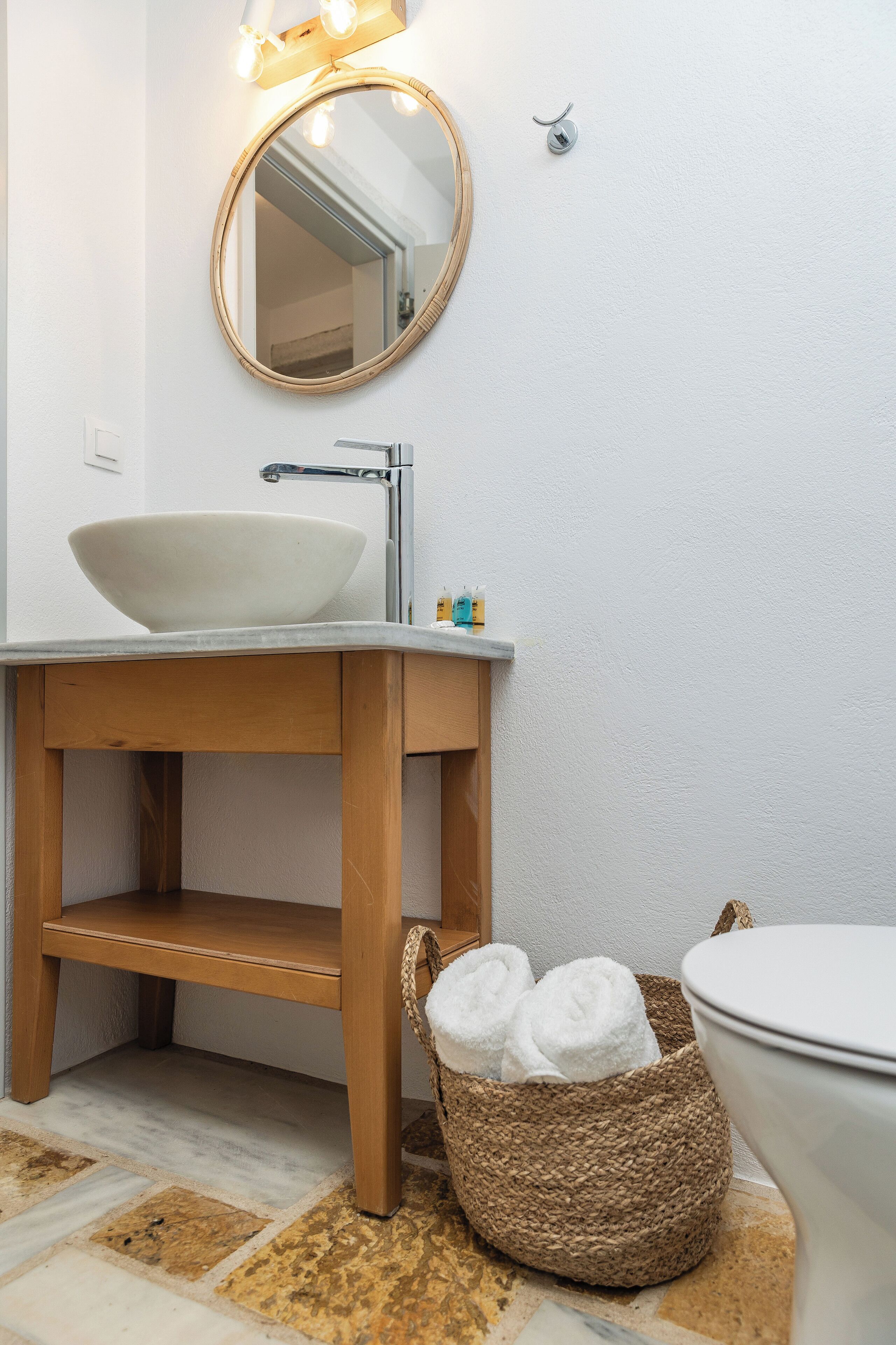 Un baño con tocador de madera, lavamanos blanco, espejo redondo y una cesta de mimbre con toallas.