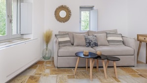 A living room with a beige sofa, two small round tables, a decorative wall hanging, and a window with a view of greenery. - Serenity Village (Naxos)