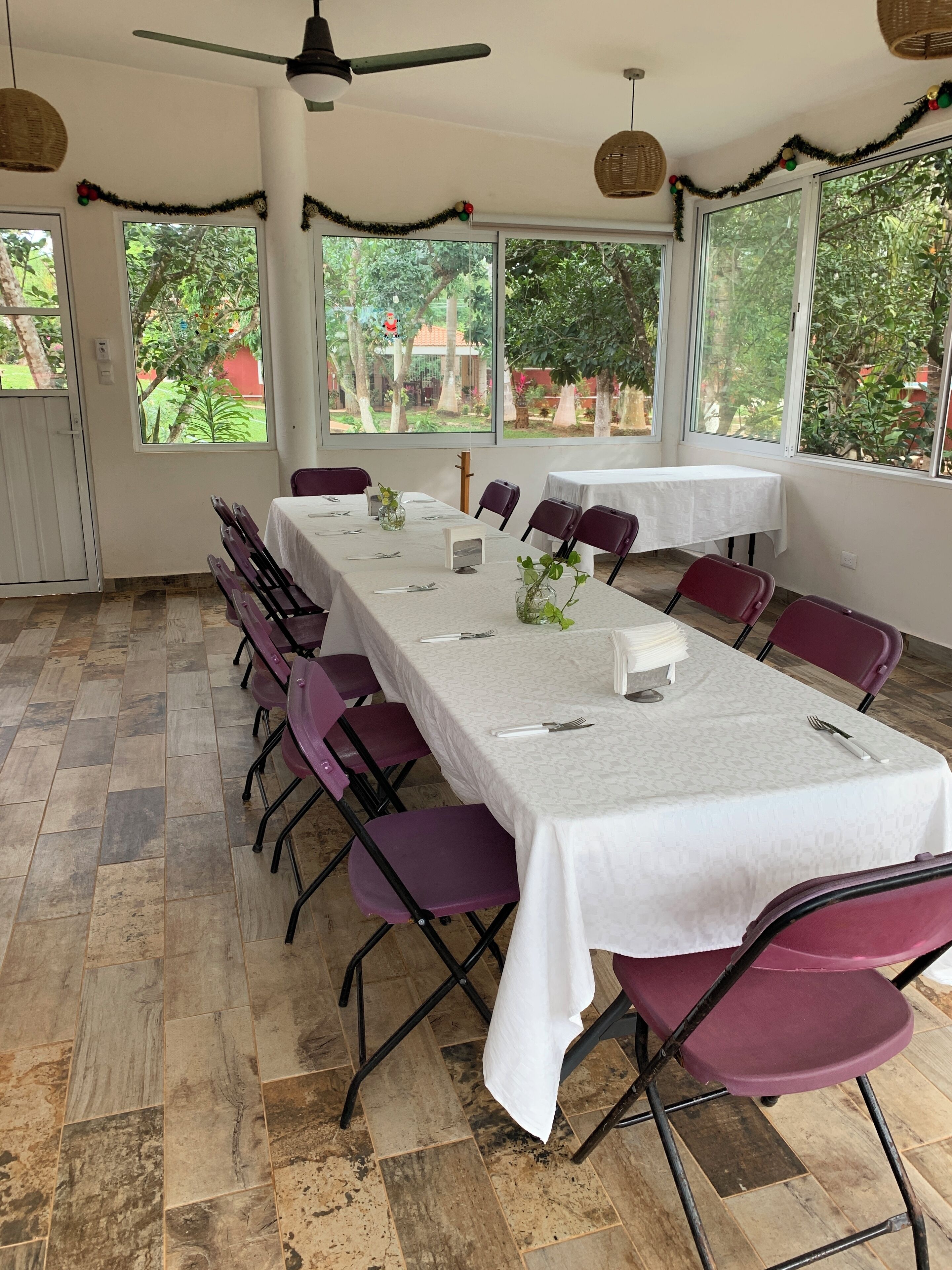 Un espace repas avec une longue table dressée pour un repas, entourée de chaises violettes, de grandes fenêtres donnant sur un paysage verdoyant et d’un ventilateur de plafond.
