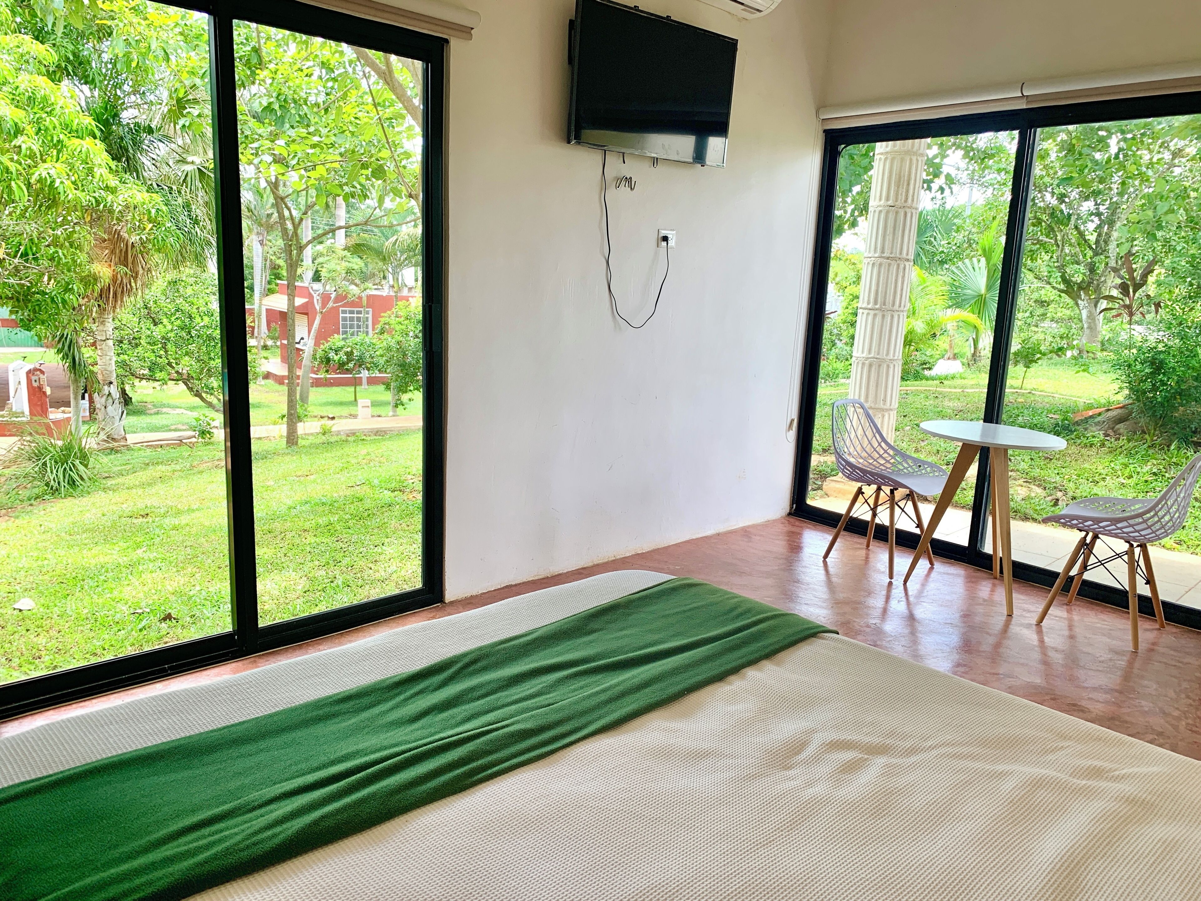 Habitación de hotel con ventanas amplias, una cama, un televisor y una pequeña mesa con sillas.