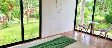Habitación de hotel con ventanas amplias, una cama, un televisor y una pequeña mesa con sillas.
