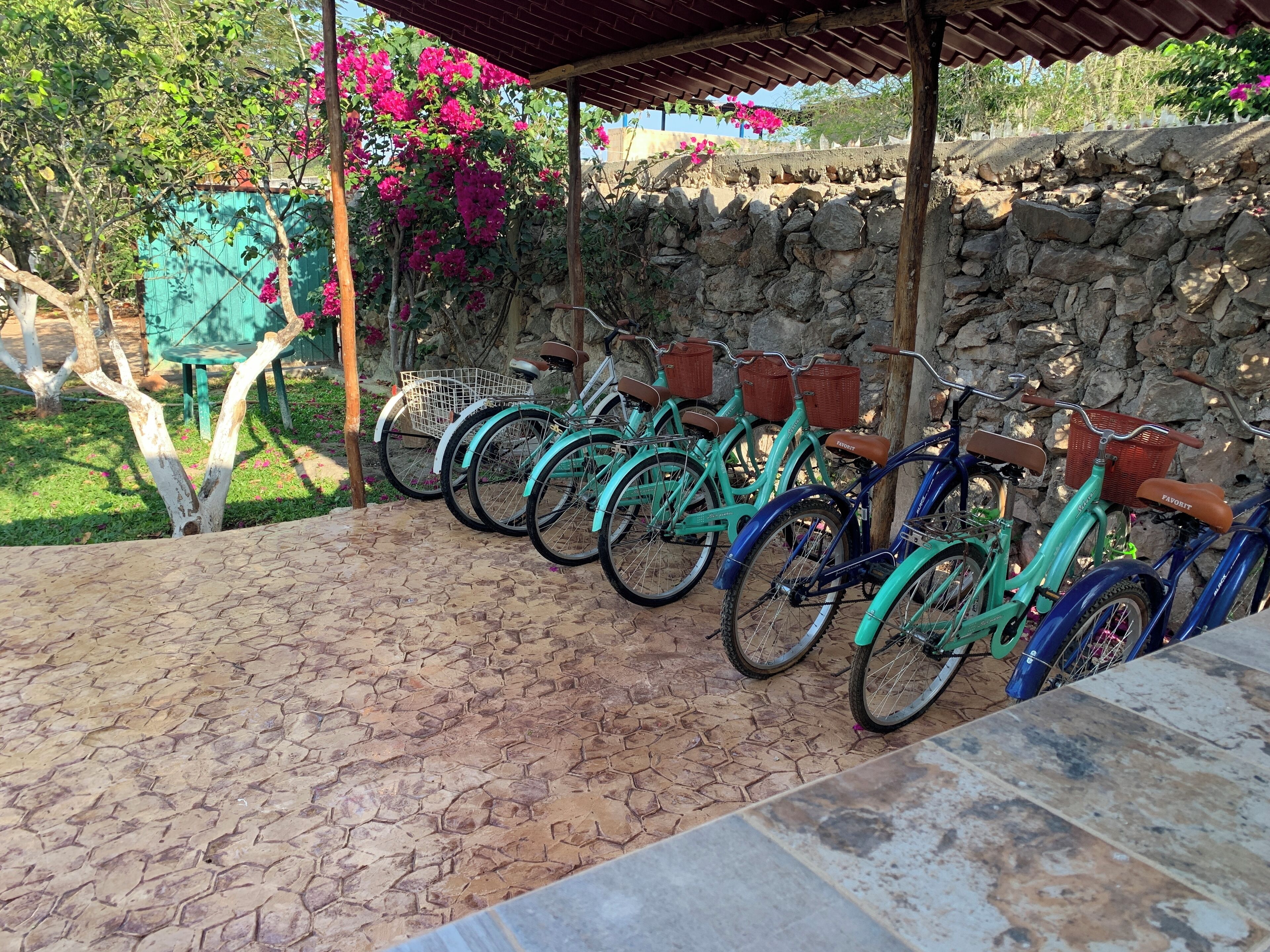 Varias bicicletas estacionadas bajo una zona techada, con un muro de piedra al fondo.