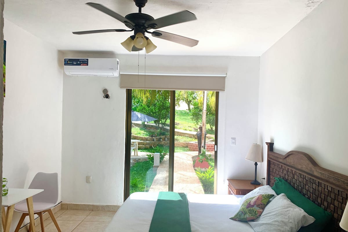 Una camera d'albergo con un letto, una ventola a soffitto, una finestra vista giardino e un tavolo con una sedia.