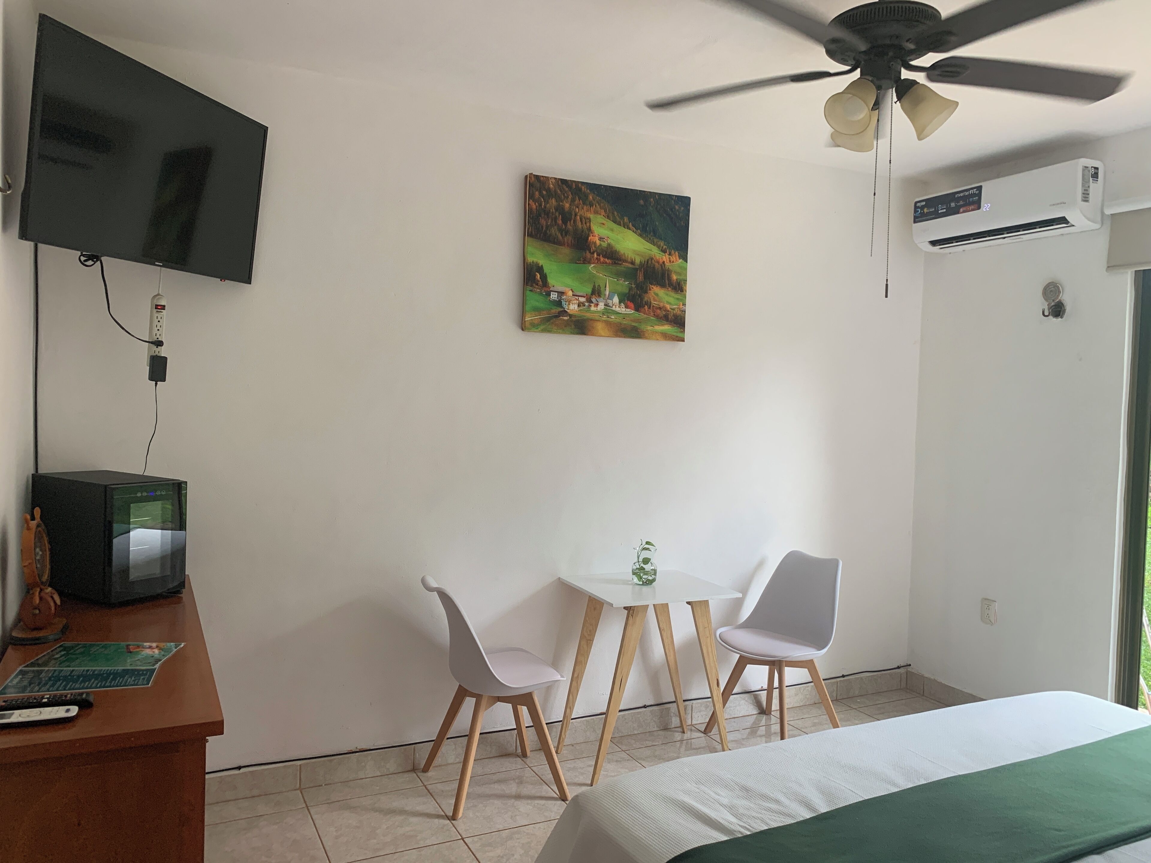 A hotel room with a bed, a desk with a computer, two chairs, a small table with a plant, a painting on the wall, a ceiling fan, and an air conditioning unit.