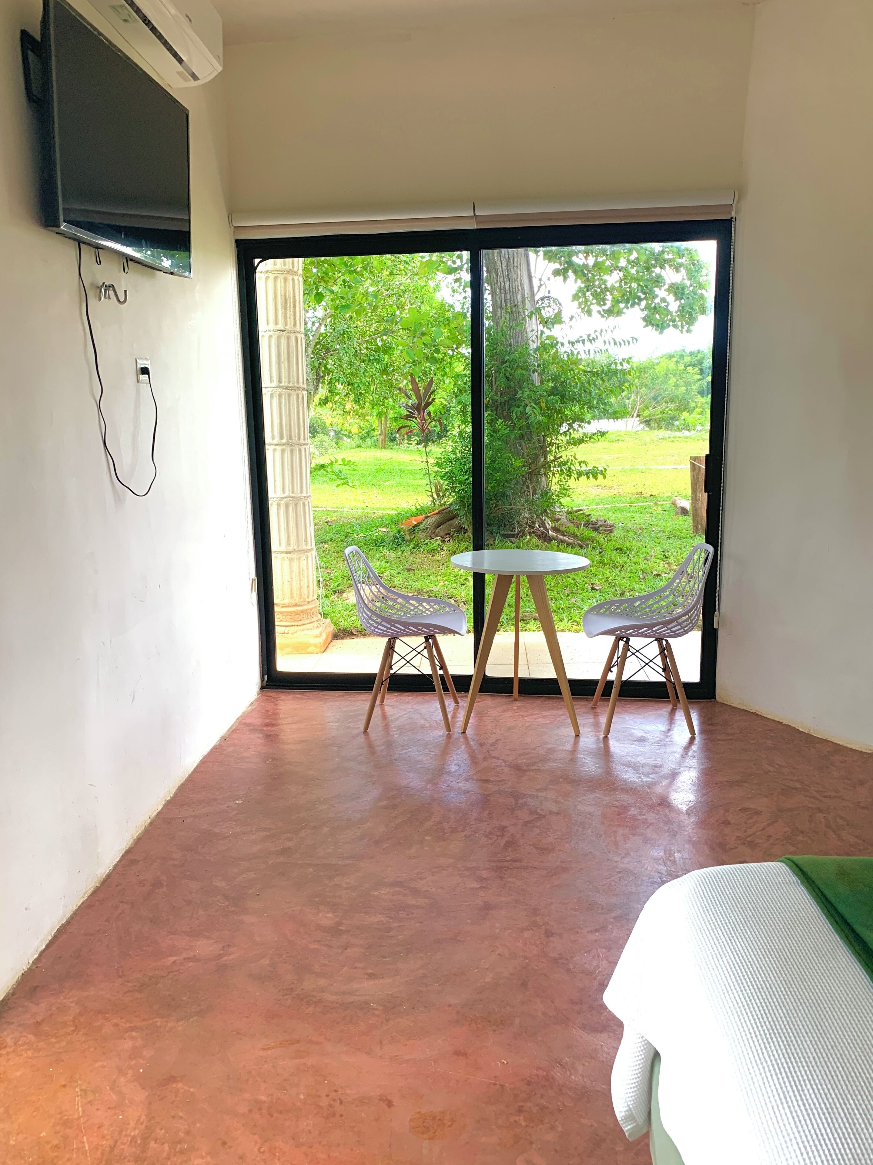 A room with a large sliding glass door leading to a garden, a small table with two chairs, and a bed with a green bedspread.