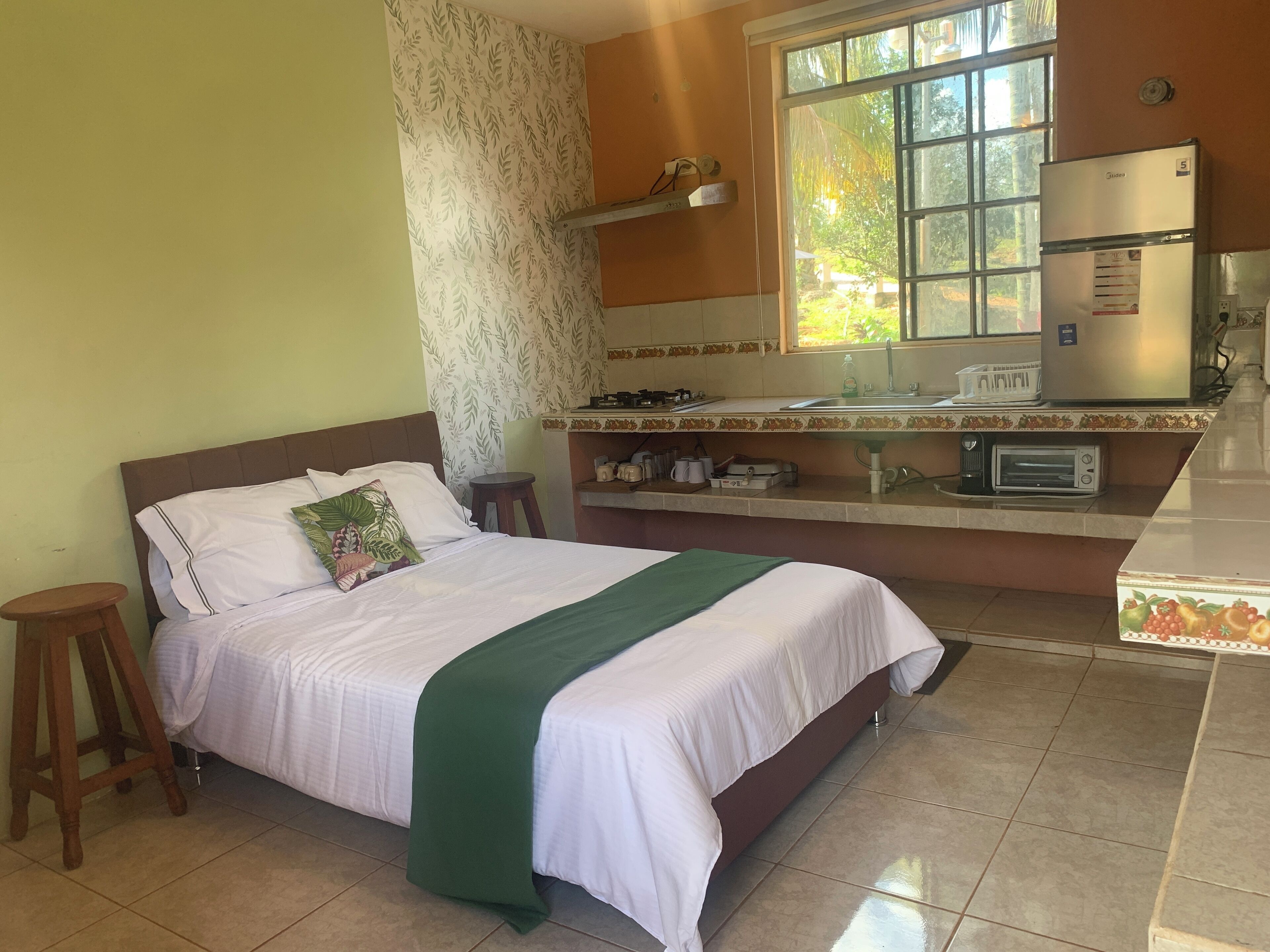 A bedroom with a bed, a green throw, a nightstand, a small stool, a window, a kitchen area with a refrigerator, a microwave, and a sink.