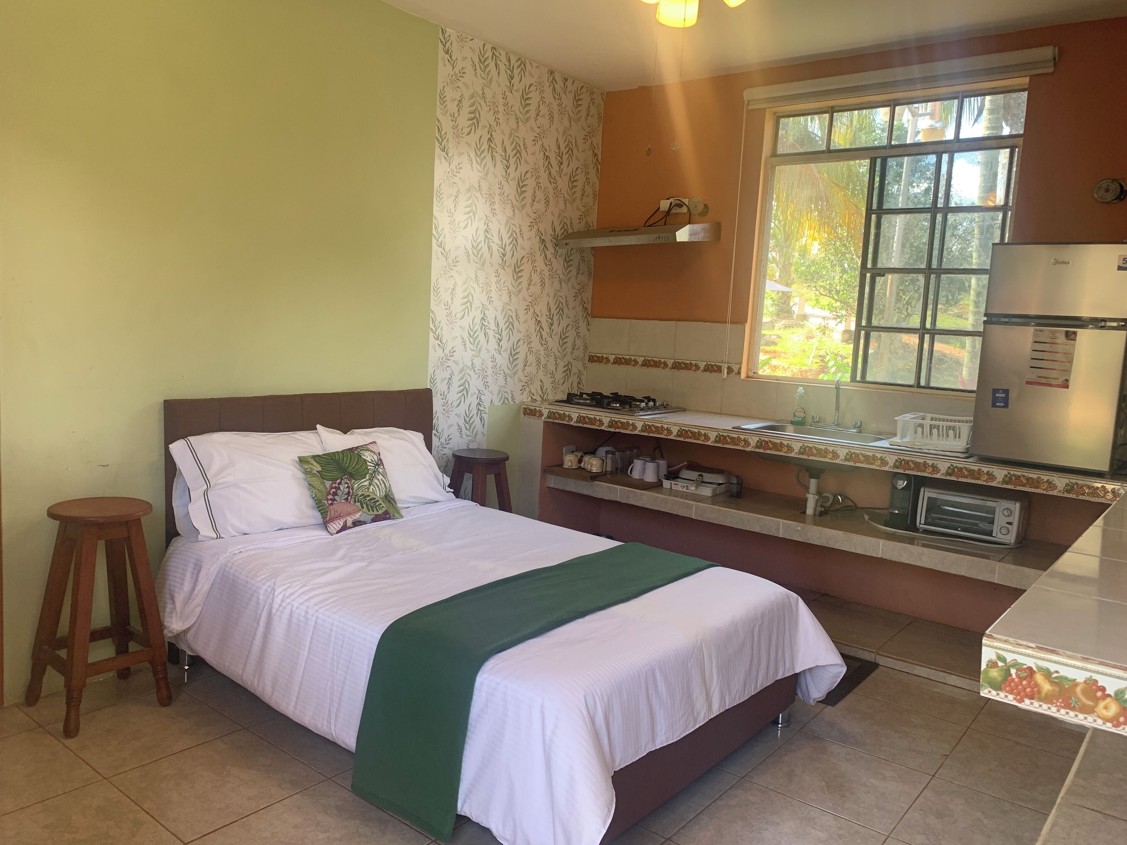 Una habitación con una cama, un taburete pequeño, una ventana con vistas a zonas verdes, una kitchenette con refrigerador y microondas.