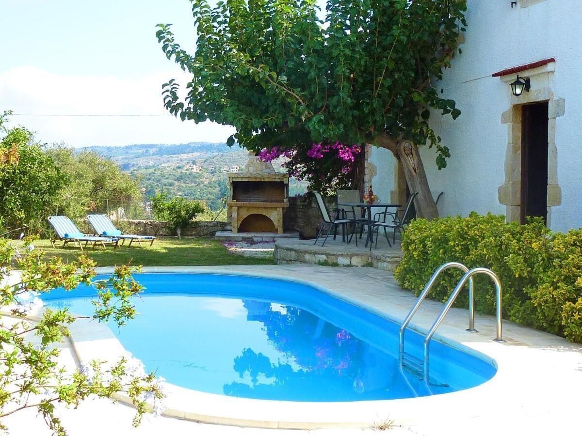 A swimming pool with a stone surround, a pergola with purple flowers, and a view of the countryside.