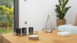 A wooden table with a tray holding three black mugs, a white tray with stacked plates, and a geometric glass candle holder. A potted plant and a bed with a light-colored blanket are in the background. - Cozy Christina Apartment ground floor (Kissamos)