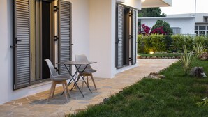 A modern building with large windows and shutters, a stone pathway, and a small outdoor seating area with a table and chairs. - Cozy Christina Apartment ground floor (Kissamos)
