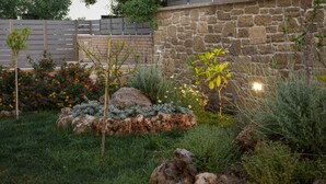 A garden with various plants and rocks, a stone wall, and a wooden fence. - Cozy Christina Apartment ground floor (Kissamos)