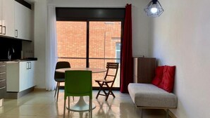 A compact living space with a kitchenette, a dining area with two chairs, a sofa with a red pillow, and a large window with a view of a brick building. - Estancias Mensuales Terrassa (Terrassa)