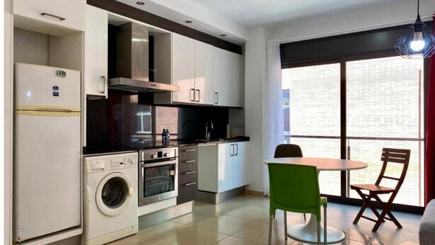 A compact kitchen with a washing machine, oven, and dining area with a table and chairs. - Estancias Mensuales Terrassa (Terrassa)
