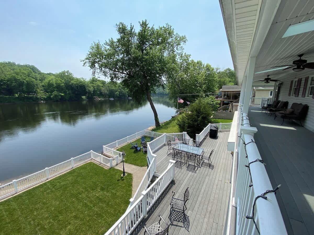 Une terrasse dotée de mobilier d’extérieur, donnant sur un lac, entourée d’une clôture blanche et d’un arbre.