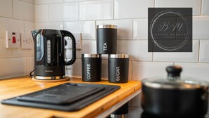 Een keuken met een zwarte koffiezetapparaat, een waterkoker en drie zwarte bakjes met de tekst thee, koffie en suiker.