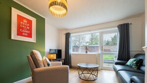 Een woonkamer met een bank, fauteuil en een kleine tafel. Op de muur hangt een ingelijste poster en er is een raam met gordijnen.