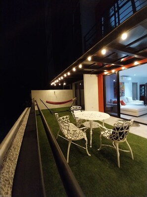 An outdoor seating area with white metal chairs and a round table, surrounded by artificial grass, under a covered section with string lights. - Luxury 3BR Family Condo in Escalón, City Views, WiFi, Parking (Apt. 102)