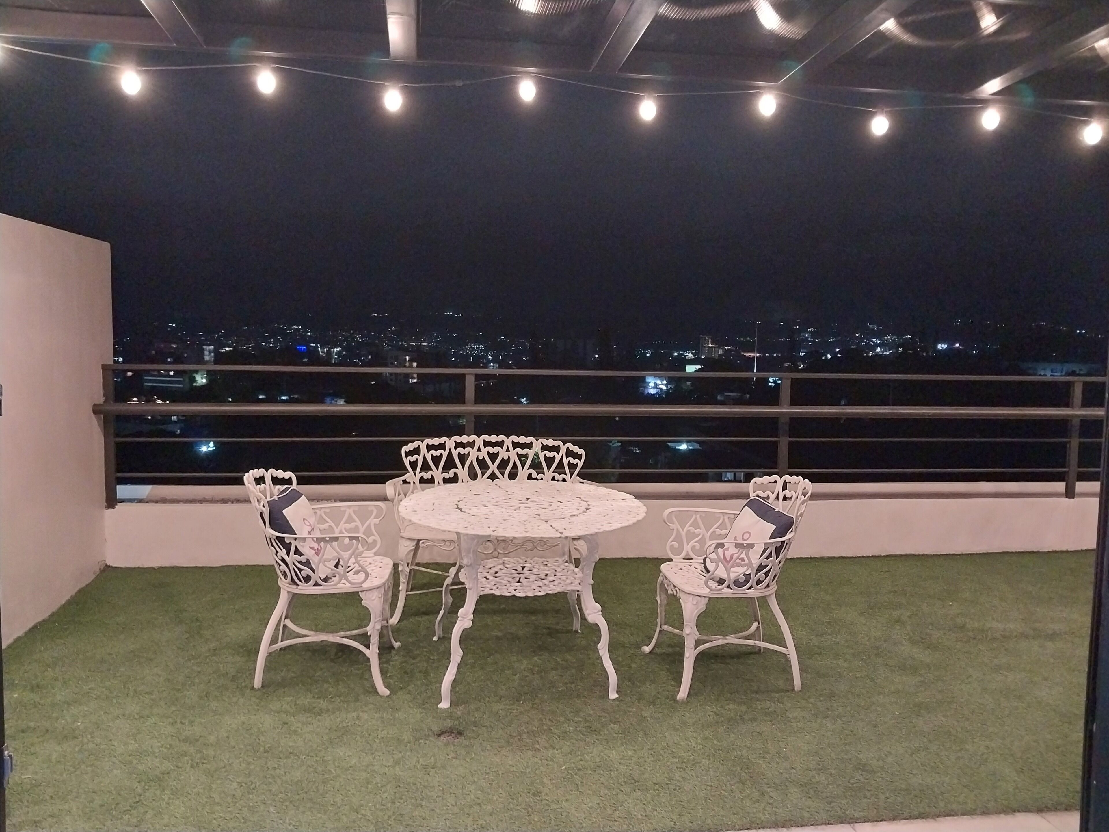 Un balcón con una mesa redonda y cuatro sillas, rodeado de césped artificial, con vistas a un paisaje urbano nocturno.