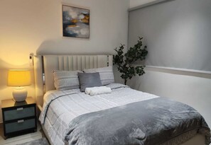 Un dormitorio con cama, mesitas de noche, una lámpara, una planta y un cuadro en la pared.