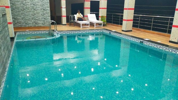 An indoor swimming pool with a tiled border, lounge chairs, and a person sitting on one of the chairs. - BLUESTONE HOTEL & RESTAURANT (Kanthi)