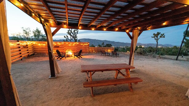An outdoor patio with a wooden table and chairs, string lights, and a view of the desert landscape. - Cozy casita on quiet desert hillside with views, fire pit and grill! (Yucca Valley)