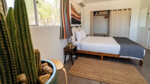 A bedroom with a bed, a nightstand, a lamp, a large window, a cactus, and a wardrobe. - Cozy casita on quiet desert hillside with views, fire pit and grill! (Yucca Valley)