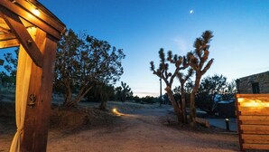 A wooden structure with a door, surrounded by desert vegetation and a dirt path leading to a building. - Cozy casita on quiet desert hillside with views, fire pit and grill! (Yucca Valley)