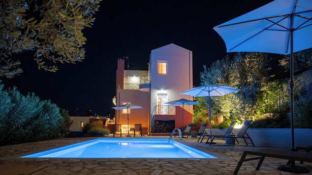 Une piscine avec des chaises longues et des parasols, une terrasse en pierre et un bâtiment doté de balcons et d’éclairages.