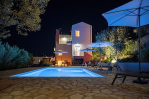 A swimming pool with lounge chairs and umbrellas, a stone patio, and a building with balconies and lights.