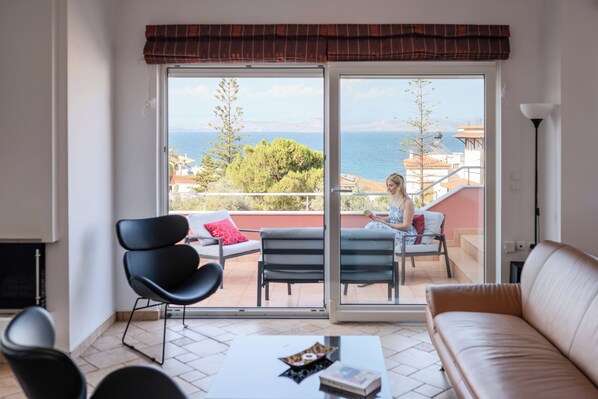 A room with a sofa, two chairs, a coffee table, and a view of the sea through a glass door.