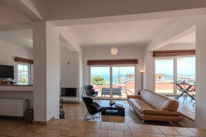 Een ruime woonkamer met een groot raam en uitzicht op zee, een bank, een salontafel en een televisie.