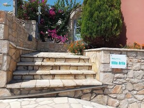 Una escalera de piedra conduce a una villa llamada Villa Artia, rodeada de vegetación exuberante y plantas con flores.