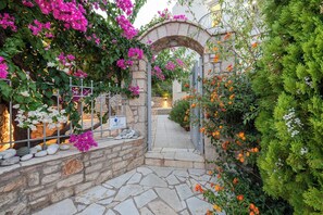 A stone archway leading to a courtyard with a stone floor, surrounded by lush greenery and flowering plants.
