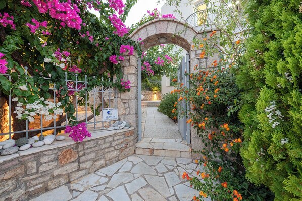 A stone archway leading to a courtyard with a stone floor, surrounded by lush greenery and flowering plants.