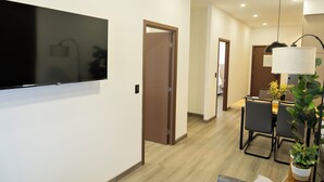 Et moderne hotelværelse med et fladskærms-tv på væggen, en trædør, et spiseområde med bord og stole, samt en potteplante.