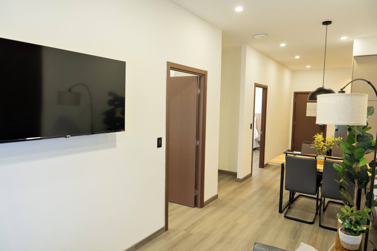 A modern hotel room with a flat-screen TV mounted on the wall, a wooden door, a dining area with a table and chairs, and a potted plant.
