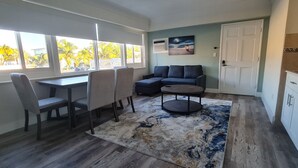 A living room with a dining area, a sofa, a coffee table, and a view of palm trees through large windows.