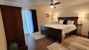 Un dormitorio con cama de madera, dos lámparas de noche, ventilador de techo y vistas al exterior a través de una ventana con cortinas.