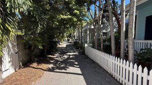 A narrow, tree-lined street with a white picket fence on one side and a row of single-story houses on the other. - Beautiful Old Town 2 bedroom Cottage on a Private Lane (Key West)