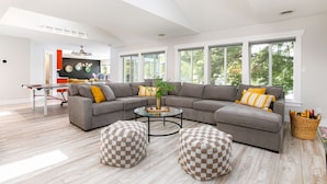 Ein modernes Wohnzimmer mit einer Eckcouch, Poufs, einem Couchtisch und großen Fenstern.