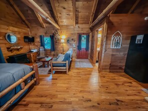 A cozy cabin interior with a wooden floor, a sofa, a chair, a small table, and a bed.