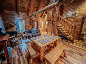 Interior acogedor de una cabaña de troncos con una escalera de madera, un sofá, una mesa de centro y una mesa de comedor con manteles individuales.