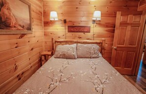 Une pièce en bois avec un lit, deux lampes fixées au mur, un tableau et une porte.