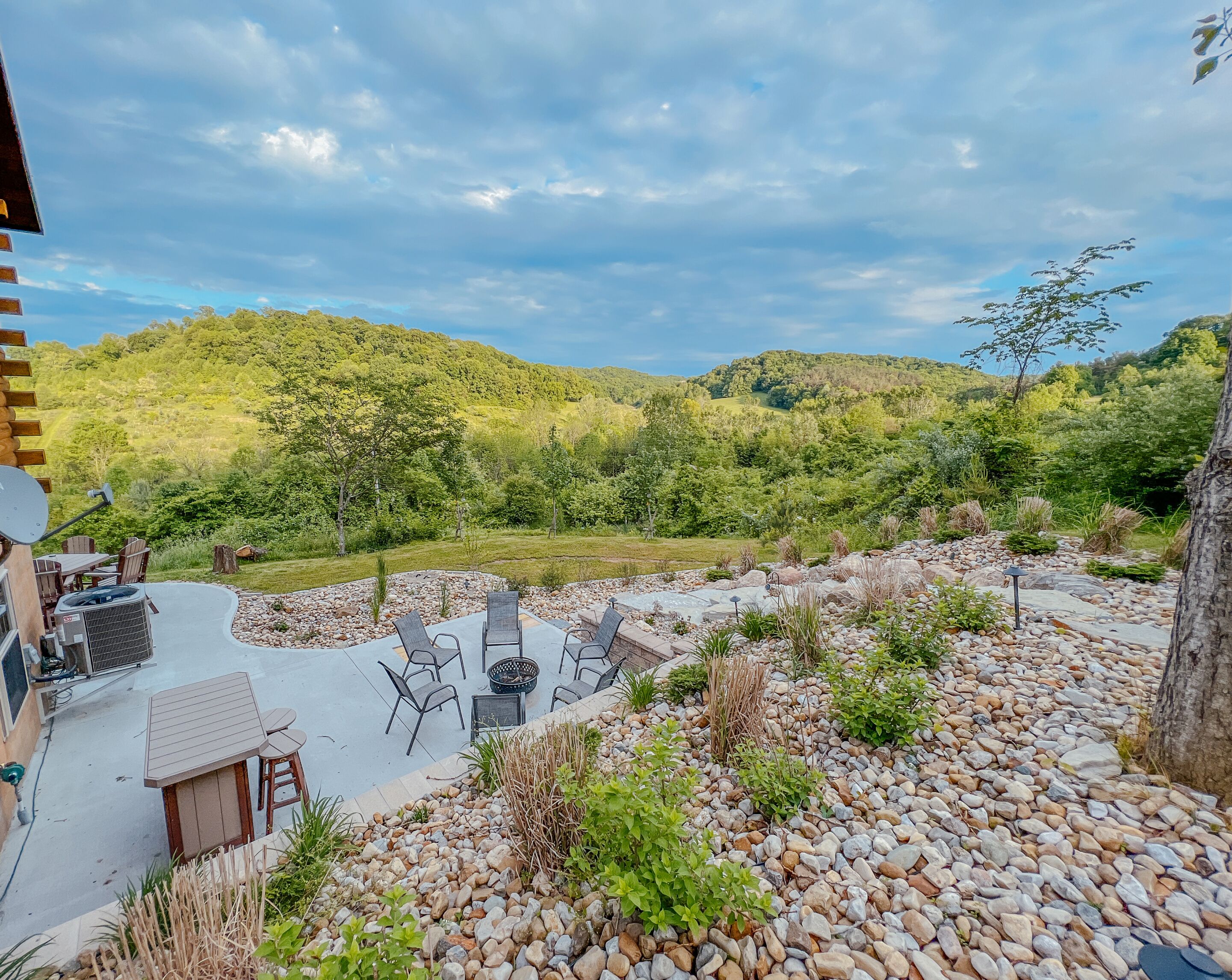 An outdoor patio with a seating area, surrounded by a garden with various plants and a view of a forested hillside.